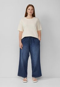 Kremowa koszulka z krótkim rękawem zestawiona z szerokimi ciemnoniebieskimi spodniami dżinsowymi i białymi sandałami. Koszulka ma okrągły dekolt; spodnie posiadają regulowany sznurek.