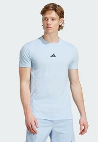 Světle modré tričko s krátkým rukávem na sportovní aktivity vyrobené z měkkého materiálu, na hrudníku s malým černým logem Adidas. Hladký povrch a přiléhavý střih.