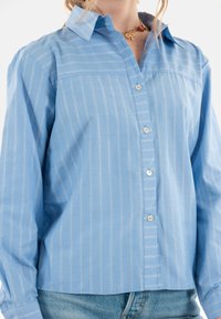 Camicia leggera azzurra con strisce verticali bianche, dotata di colletto e maniche lunghe, realizzata in tessuto morbido. Chiusura semplice con bottoni.