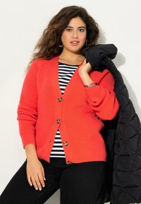 Korallfärgad stickad cardigan med V-hals, fyra knappstängningar och ribbad textur, bärs över en svartvit randig topp, med en svart kappa.