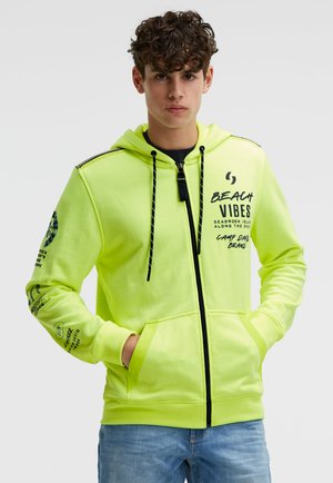 Junger Mann mit lockigem Haar trägt einen neon-grünen Kapuzenpullover mit Reißverschluss und schwarzem Schriftzug "Beach Vibes" sowie blaue Jeans, Hände in den Taschen, neutraler Hintergrund.