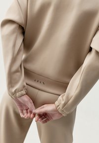 Beige överdimensionerad sweatshirt med elastiska manschetter, i kombination med matchande byxor. Har en subtil logodetalj vid midjan. Slät textur.