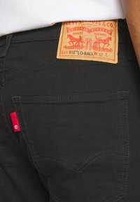 Svarta jeans i denim med en gul läderpatch baktill som har röda grafikdetaljer, en liten röd etikett på sidan och kontrasterande sömmar.