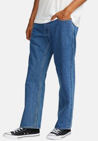 Jeans en denim bleu avec une coupe décontractée, présentant une texture décolorée et un design à cinq poches. Associés à des baskets en toile noires.