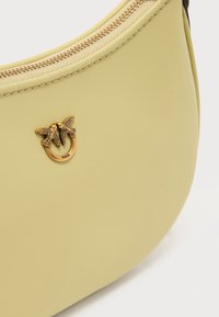 Κίτρινη δερμάτινη τσάντα με στρογγυλεμένο σχήμα, με μεταλλικό λογότυπο σε χρυσό τόνο με δύο πουλιά, κλείσιμο με φερμουάρ και εμφανείς ραφές.