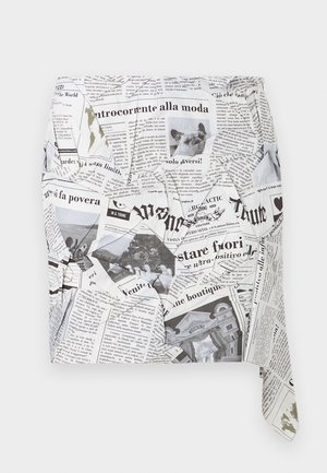 Žena s tmavšou pleťou, oblečená v čiernom asymetrickom minišati s potlačou novín a čiernych lodičkách so zapínaním na členku, stojaca s jednou rukou v vrecku.