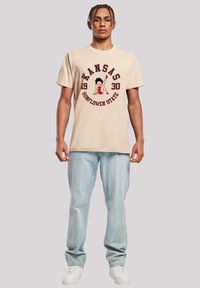 Beige cotton t-shirt with Kansas graphic, featuring a playful design. Paired with light blue denim jeans and white sneakers.