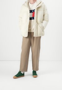 Bílá péřová bunda s kapucí, béžový pletený svetr s barevným logem a světle hnědé kalhoty. Zelené tenisky s hnědými podrážkami.