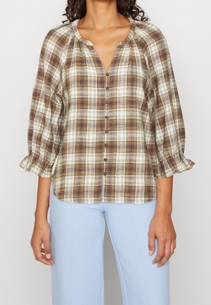 Femme portant un chemisier à carreaux marron et crème boutonné avec des manches trois-quarts volantées et un pantalon bleu clair.