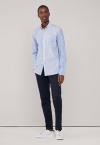Blau-weiß gestreifte Hemd mit Knöpfen, lange Ärmel, eine Brusttasche, kombiniert mit dunklen Hosen und weißen Schuhen.