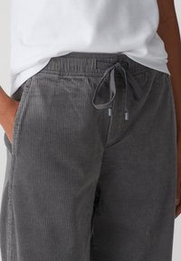 Pantalon en velours côtelé gris avec taille élastique et cordon de serrage. Comprend des poches latérales et une texture côtelée. Porté avec un t-shirt blanc.