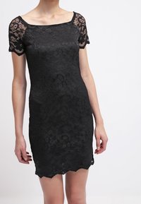 Černé krajkové šaty s krátkými květinovými rukávy, přiléhavým střihem a vlnitým lemem. Texturovaný materiál dodává hloubku.