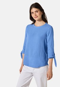 Top in cotone blu chiaro, testurizzato, con maniche a tre quarti, dettagli con polsini annodati e scollatura rotonda, abbinato a pantaloni bianchi.