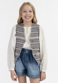 Woven vest z geometrycznym wzorem w niebieskim i kremowym kolorze, z złotymi akcentami, nałożony na białą bluzkę i zestawiony z szortami dżinsowymi.