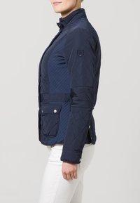 Granatowa pikowana kurtka z wysokim kołnierzem, bocznymi kieszeniami i ozdobnym przeszyciem. Posiada naszywkę z logo na lewym rękawie oraz zatrzaski jako dodatki.