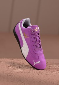 Violetti nubukistani urheilusandaalit valkoisilla yksityiskohdilla, joissa on teksturoitu muotoilu ja kultainen Puma-logo, pyöristetty kärki ja musta pohja.