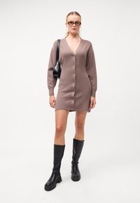 Pletené šaty v taupe barvě s výstřihem do V, dlouhými rukávy, předními knoflíky a bočním rozparkem, doplněné černými vysokými kozačkami a černou kabelkou.