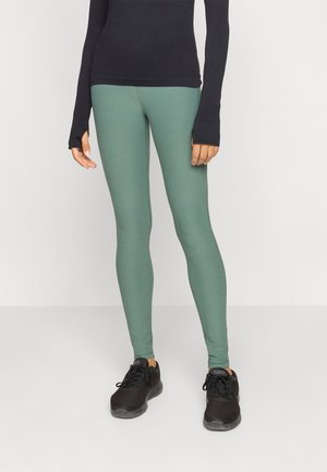 Persona con leggings verdi, maglietta a maniche lunghe nera e scarpe da ginnastica nere, in piedi su un pavimento chiaro contro una parete chiara.