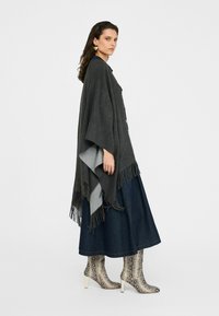 Grijze wollen poncho met franjes, over een donkere jeansrok en slangenprint enkellaarsjes met een hoge conische hak.