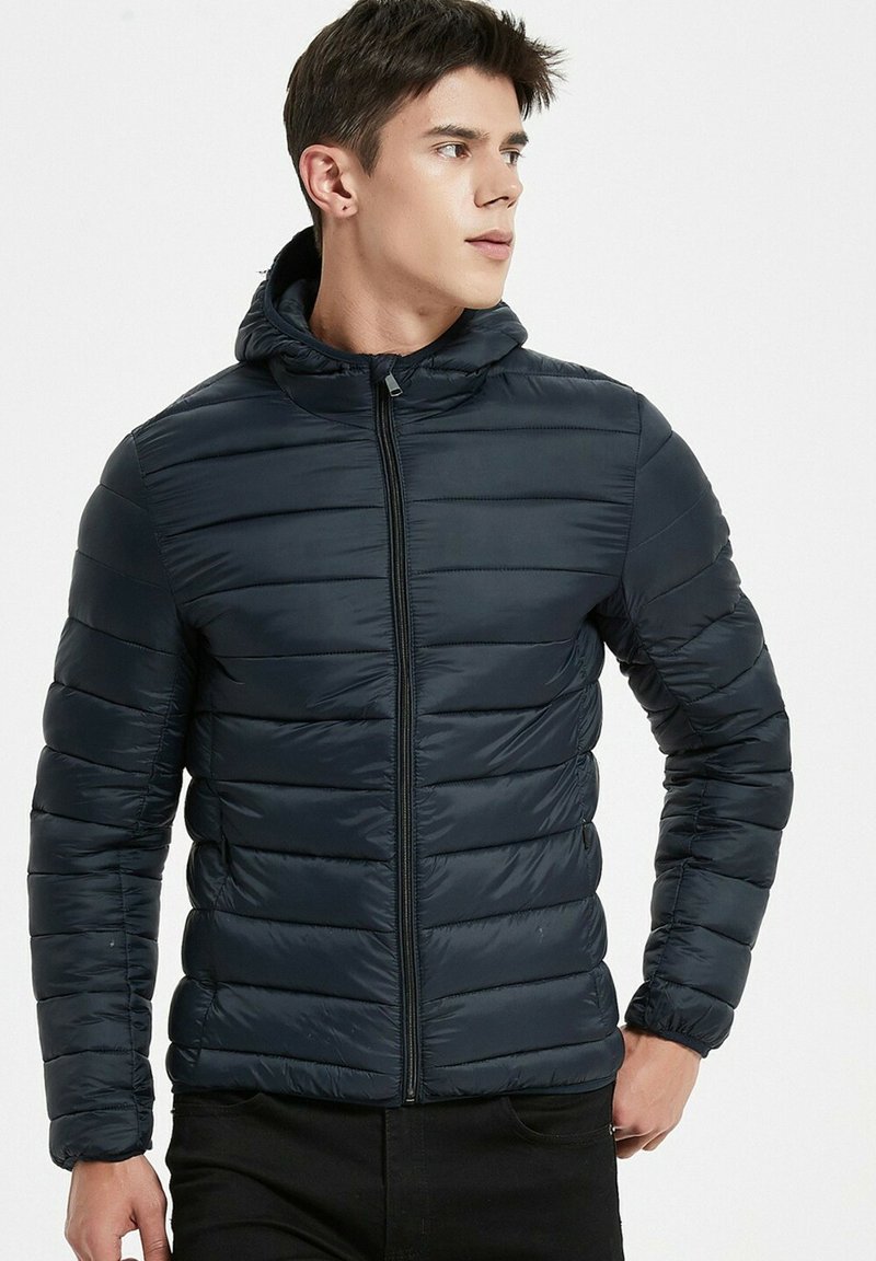 Veste matelassée bleu marine avec capuche, dotée d'un design matelassé horizontal, d'une fermeture éclair et de poignets élastiques pour un ajustement confortable.