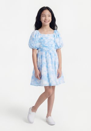 Niña joven con cabello largo y oscuro, vestida con un vestido azul y blanco de mangas abullonadas y zapatos blancos sin cordones, de pie con las piernas cruzadas y sonriendo.