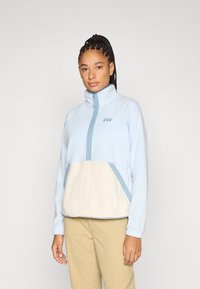 Ανοιχτό μπλε φλις πουλόβερ με φερμουάρ στο μισό μήκος, ψηλό γιακά, μπεζ μπροστινή επιφάνεια και ανοιχτό μπλε λεπτομέρειες. Διαθέτει κεντητό λογότυπο στο στήθος.