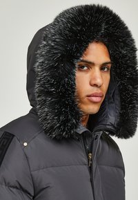 Dunkelgraue Puffjacke mit abnehmbarem schwarzen Futter, strukturierten Akzenten, Reißverschluss und Logodetail auf der Schulter.