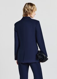 Persona con capelli biondi corti in piedi di spalle, vestita con un completo blu navy e che tiene una pochette in pelle nera nella mano destra.