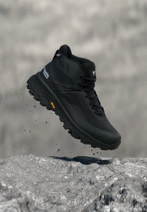 Czarny but trekkingowy Columbia z podeszwą Vibram unoszący się nad skalistym terenem, cząsteczki kurzu unoszące się z miejsca uderzenia.