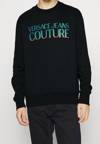 Svart sweatshirt med texten "VERSACE JEANS COUTURE" i iridescerande bokstäver, rundad halsringning, långa ärmar och ribbade muddarna.