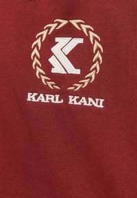 Tessuto rosso con logo "K" ricamato in bianco circondato da una corona di alloro beige e il testo "KARL KANI" sottostante.