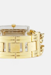 Guldmetallur med et rektangulært, gennemsigtigt urkasse. Remmen har sammenkoblingselementer, accentueret med indgraverede detaljer.