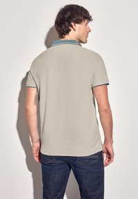 Grå pikétröja i en texturerad tygtyp, med en kontrasterande teal-färgad krage och ärmslut. Kombineras med mörkblå jeans.