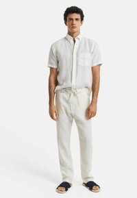 Linnen shirt met korte mouwen en joggers in gebroken wit, gecombineerd met marineblauwe slippers. Fris, licht materiaal met een zakdetail.