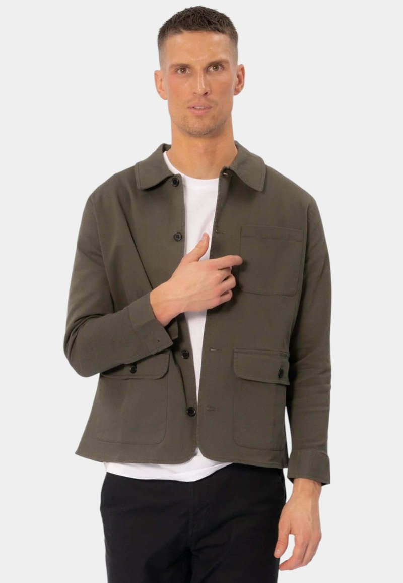 Oliven grøn jakke med krave, frontknaplukning, to brystlommer og en hvid skjorte nedenunder. Har en afslappet pasform og lange ærmer.
