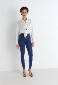 Vit långärmad skjorta knuten i midjan, i kombination med mörkblå skinny jeans och svarta remsandaler med klack. Enkel, figursydd design.