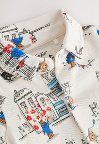 Krekls ar pogām no teksturēta krēmkrāsas auduma, rotāts ar krāsainām ilustrācijām ar ēkām un tēliem, akcentēts ar koka pogām.