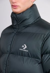 Ciemnozielona pikowana kurtka z wysokim kołnierzem, przednim zamkiem błyskawicznym oraz haftowanym logo "Converse" na lewej piersi. Gładka, puchowa tekstura.
