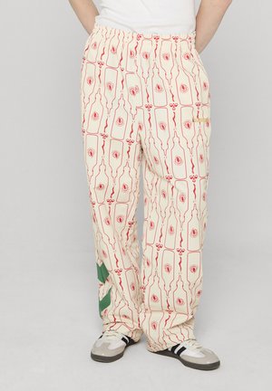 Persona con pantaloni color crema, bottiglia rossa e motivi floreali, striscia verde vicino alla caviglia, sneakers bianche con strisce nere.