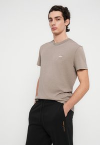 Ανοιχτό καφέ βαμβακερό t-shirt με κοντά μανίκια και στρογγυλή λαιμόκοψη. Διαθέτει μικρό λογότυπο στο στήθος. Φοριέται με μαύρες φόρμες.