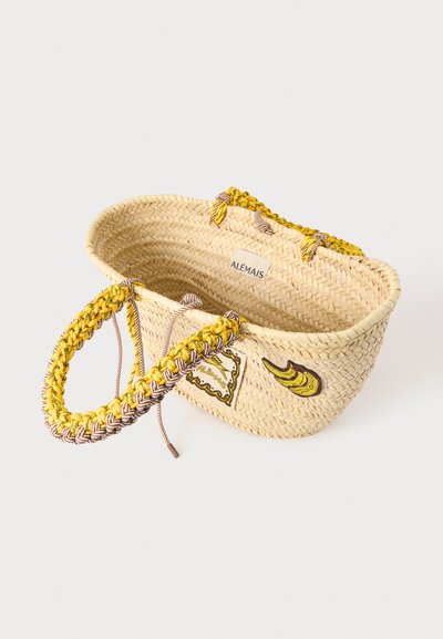 Sac cabas en paille tressée avec des anses tressées jaunes et marron, décoré d'un patch banane et d'une étiquette de marque portant la mention "ALÉMAIS" à l'intérieur.