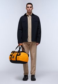Chaqueta negra con dos bolsillos en el pecho sobre una camisa beige, combinada con pantalones beige y zapatos negros; sosteniendo una bolsa de deporte negra y naranja.