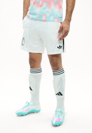 Jugador de fútbol masculino con pantalones cortos Adidas blancos, calcetas blancas hasta la rodilla con rayas, espinilleras y tacos azul claro con detalles rosas.