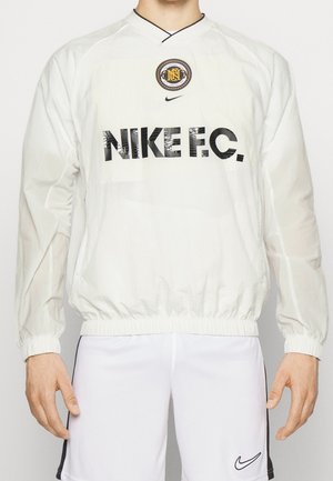Osoba nosząca biały sportowy pulower Nike F.C. z czarnym logo i emblematem, połączony z białymi szortami z czarnym znakiem Nike swoosh.
