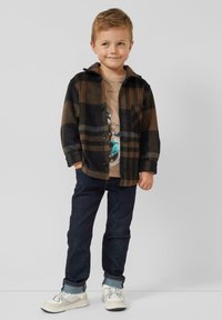 Geruit flanellen overhemd in zwart en bruin, effen beige T-shirt met opdruk, donkere spijkerbroek en lichte sneakers met grijze en witte accenten.