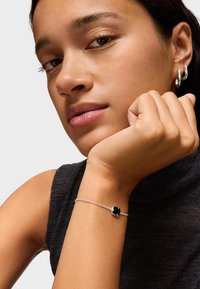 Pulsera de cadena de plata con un charm en forma de flor negra. El modelo lleva un cuello de tortuga oscuro, con un tono de piel suave y natural.