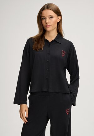 Junge Frau mit glatten braunen Haaren, die ein schwarzes Hemd zum Knöpfen und dazu passende Hosen mit roten "JOOP! JEANS" Logos trägt.