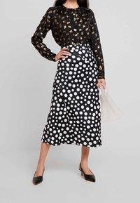 Blusa nera con stampa astratta dorata, abbinata a una gonna midi a pois neri e bianchi. La modella tiene in mano una borsa trasparente con perline.