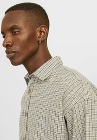 Chemise à carreaux beige avec un col button-down, présentant un motif de grille avec des lignes bleu foncé et une texture légère. Détail subtil de boutons à l'avant.