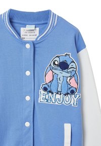 Giacca varsity blu chiaro con maniche bianche. Presenta un design ricamato testurizzato di un personaggio e la parola "ENJOY" in blu.